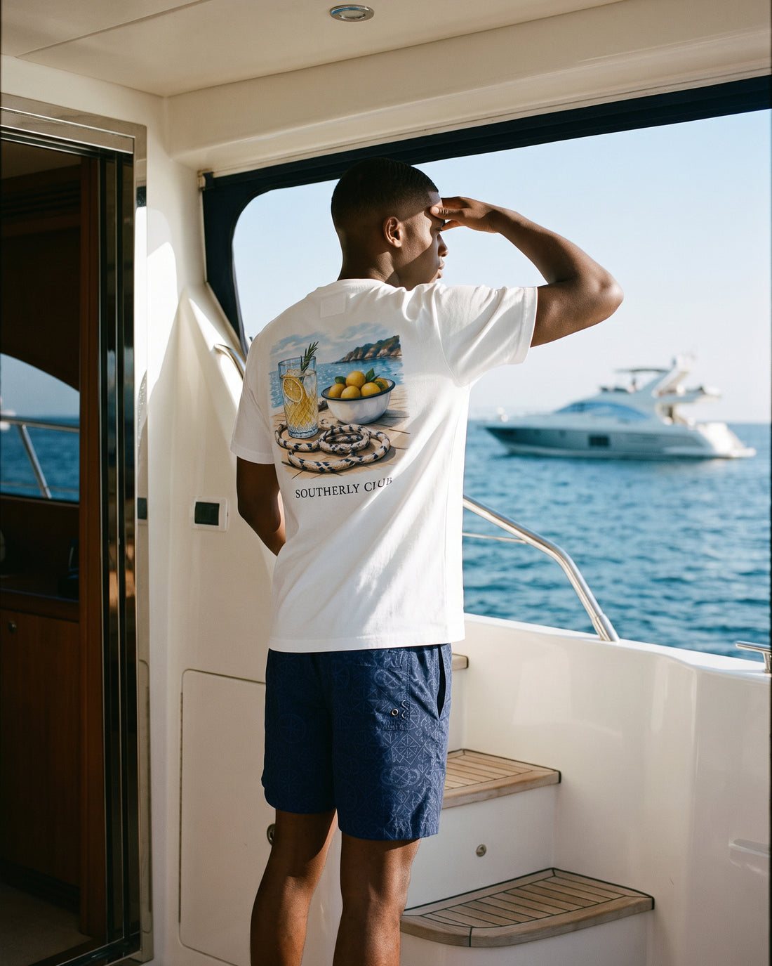 Campaign concept: man wearing a white citrus graphic tee and navy shorts on a yacht. Garment details and fit are unconfirmed.
