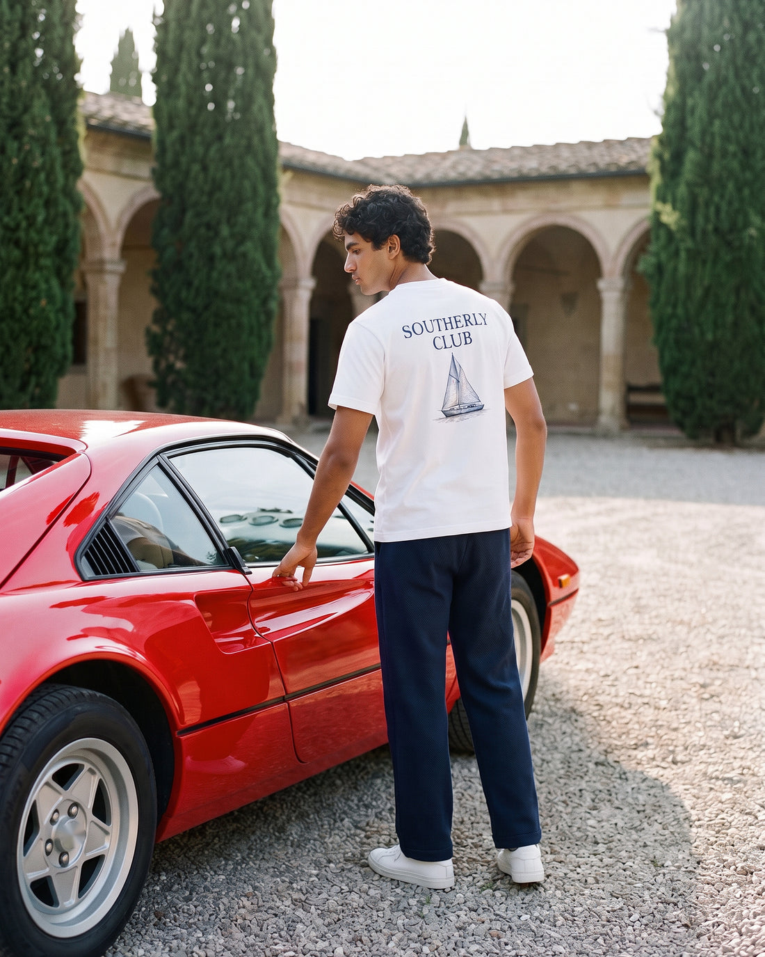 Campaign concept: man wearing a white sailboat graphic tee beside a red car. Garment details and fit are unconfirmed.