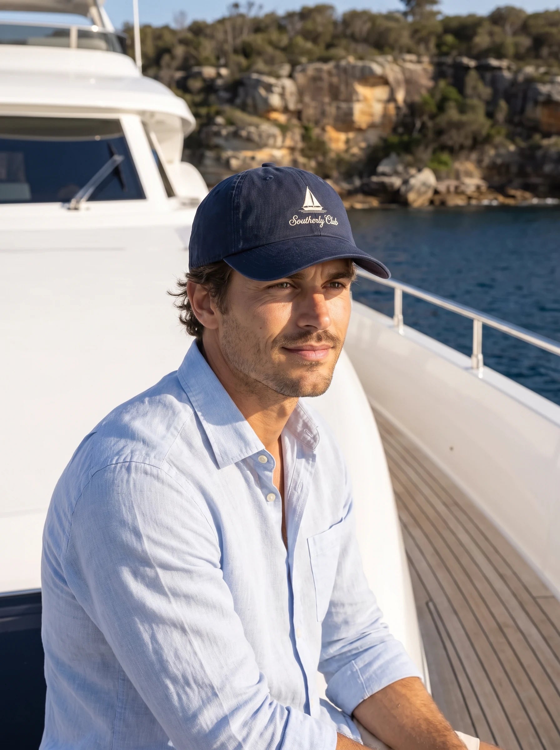 AI-generated campaign concept: Close-up of an adult model wearing the navy sailing cap with cream boat embroidery on a superyacht. Garment fit unconfirmed.